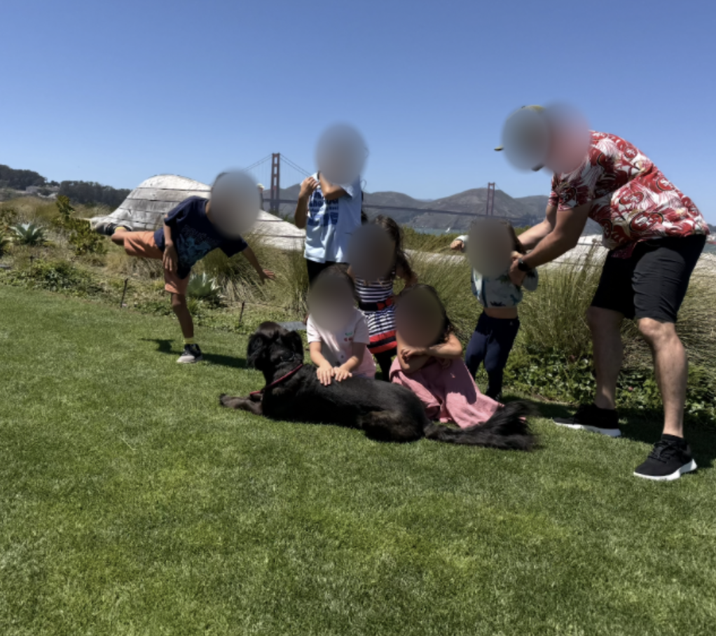 Kuma lying calmly on grass surrounded by children and their families, whose faces are blurred for privacy